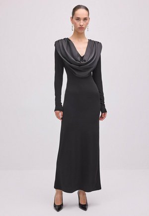 Femme portant une longue robe noire avec un col bénitier drapé, des manches longues, des talons noirs pointus et des boucles d'oreilles pendantes sur un fond uni.