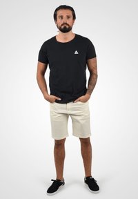 Svart bomull T-skjorte med en liten hvit trekantlogo, kombinert med kremfargede shorts og svarte sneakers. Avslappet passform med rene linjer.