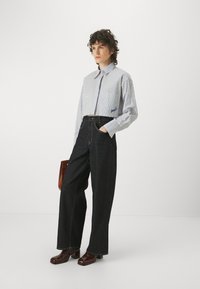 Camicia a righe, con bottoni, bianca e blu dal design cropped, abbinata a pantaloni larghi in denim scuro a vita alta e scarpe col tacco marroni.