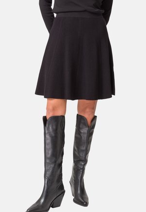 Femme portant une jupe noire évasée associée à des bottes en cuir noires montantes avec des bouts pointus et des talons compensés.