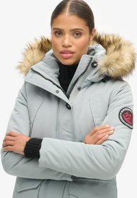 Parka azzurro chiaro con cappuccio in pelliccia sintetica, polsini in maglia nera, chiusure a zip e bottoni, e una toppa con logo sulla manica. Tessuto liscio e design imbottito.