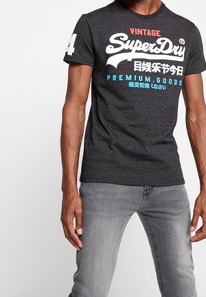 Mężczyzna ubrany w ciemnoszarą, vintage koszulkę SuperDry z białym, czerwonym i niebieskim napisem, w połączeniu z jasnoszarymi jeansami, stojący z rozluźnionymi rękami.