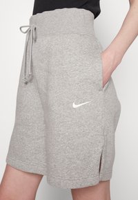 Šedé sportovní šortky vyrobené z měkké tkaniny, s elastickým pasem na šňůrku, bočními kapsami a malým bílým logem na levé straně.