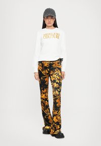 Camicia bianca a maniche lunghe con stampa dorata "Versace Jeans Couture", abbinata a pantaloni a zampa d'elefante neri e dorati e scarpe con plateau nere.