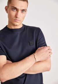 Tricou bleu marin cu mânecă scurtă, având un patch texturat în formă de pătrat pe piept, asociat cu o brățară din lanț auriu pe un braț gol.