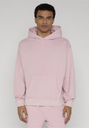 Jeune homme aux cheveux courts et bouclés portant un sweat à capuche rose clair et un pantalon assorti, debout devant un fond gris uni.