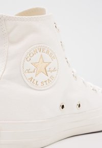 Zapatilla alta de lona blanca con parche de logo dorado, punta redonda, ojales de metal y suela de goma. Tela texturizada con detalles de costuras visibles.