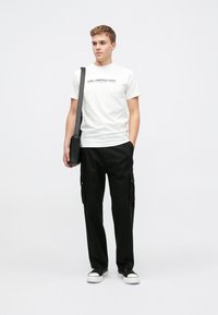 Balta T-kreklu ar melnu "Karl Lagerfeld Jeans" uzrakstu, kas apvienots ar melnām kravas biksēm ar sānu kabatām un melnām kedām ar baltām zole.