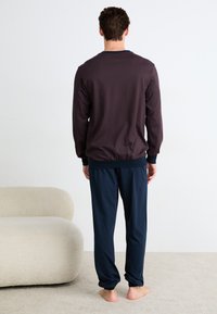 Haut long à manches dans un maron foncé avec un motif en losange, associé à un pantalon bleu marine. Les deux sont fabriqués en tissu doux et léger.