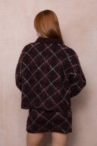 Mujer con cabello largo y rojo que lleva un suéter de lana morado oscuro y una falda a juego con un patrón de diamantes blancos, de espaldas contra una pared clara.