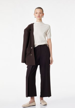 Vrouw draagt een wit kortemouwen topje, zwarte wijde broek, beige loafers en een zwarte blazer over één schouder gedrapeerd.