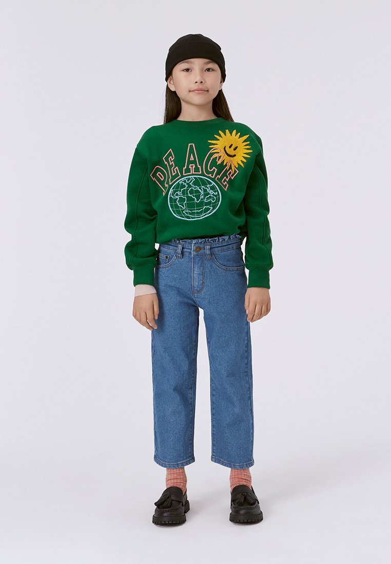 Felpa verde con testo "PEACE" e grafica del sole, abbinata a jeans blu. Scarpe nere e un berretto nero completano l'outfit.