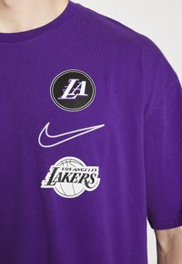 camiseta de algodón morada con tres logotipos: "LA", el swoosh de Nike y el diseño de baloncesto de "Los Angeles Lakers", todos en colores contrastantes.