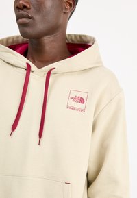 Sudadera beige con cordones y bolsillo en color burdeos; presenta un logo impreso en rosa en el lado izquierdo del pecho, tejido texturizado y corte relajado.