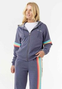 Vrouw in een blauwe hoodie met rits en bijpassende broek met regenboogstrepen op de mouwen en broekspijpen, staand met één hand in de zak.
