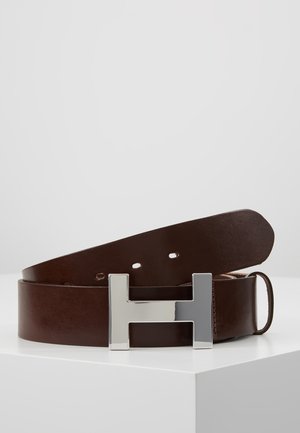 Hnědý kožený pásek s velkou stříbrnou kovovou přezkou ve tvaru písmene "H" položený na bílém rohovém povrchu.