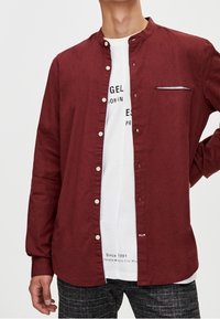 Chemise bordeaux à boutons avec col mandarin, manches longues, poche avant avec bordure blanche contrastante, portée avec un T-shirt blanc à motif en dessous.