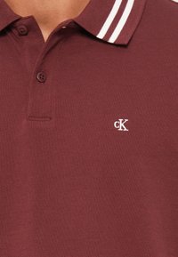 Bordó polo ing két fehér csíkkal a galléron és egy kis fehér hímzett "cK" logóval a mellkason.