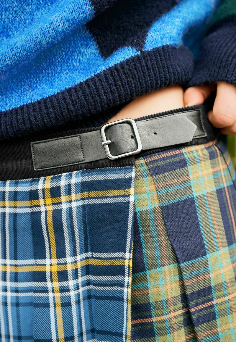 Detailní záběr na ruce osoby, které upravují černý pásek na vícebarevné kárované sukni, s modrým vzorovaným svetrem viditelným v pozadí.