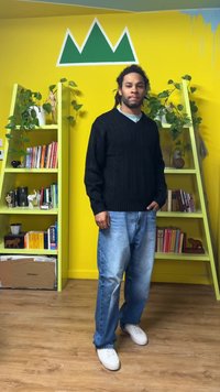 Maglione nero lavorato a maglia, camicia azzurro chiaro, jeans blu e sneakers bianche. Scaffali con piante verdi, parete gialla con grafica di montagna verde.