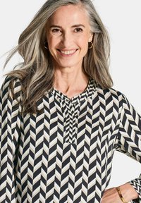 Blouse noir et blanc avec un motif en chevron, dotée d'un col rond et de détails de boutons. Fabriquée dans un tissu lisse.