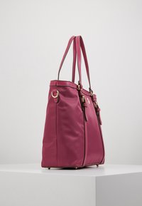 Ciemnoróżowa torba na ramię z podwójnymi paskami, złotymi klamrami i białymi przeszyciami, wyeksponowana na białym postumencie na tle jednolitego tła.