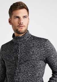 Giacca a zip grigia e nera in maglia con colletto alto, caratterizzata da un motivo testurizzato e una tasca sul petto. Tessuto morbido e taglio moderno.