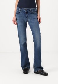 Jean en denim bleu avec une coupe droite, cinq poches et un léger dégradé. Portés avec des chaussures noires, montrant un look décontracté.