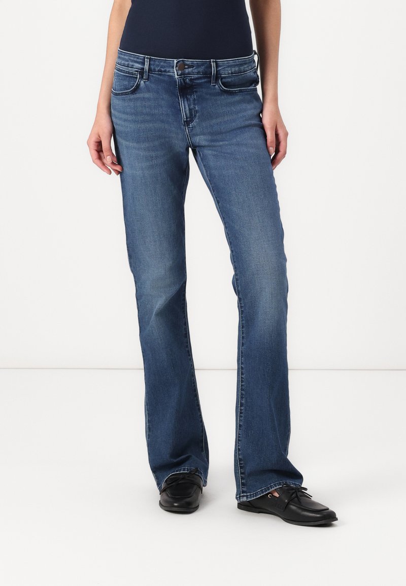 Jean en denim bleu avec une coupe droite, cinq poches et un léger dégradé. Portés avec des chaussures noires, montrant un look décontracté.