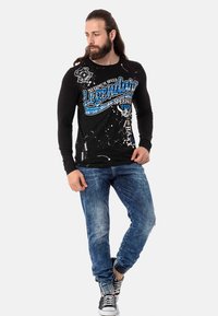 Schwarz langärmliges T-Shirt mit blauem Grafikdruck, kombiniert mit hellblauen, verwaschenen Jeans und schwarz-weißen Sneakern. Lässiger Stil.