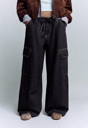 Person iført sorte cargobukser med vide ben og snøre, brun ruskindsjakke og beige ballet flats, hænderne puttet i lommerne.