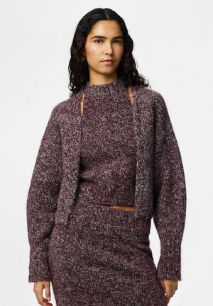 Donna con capelli ricci scuri che indossa un maglione a maglia color marrone scuro e grigio con dettagli ritagliati e gonna a maglia abbinata, su sfondo bianco.