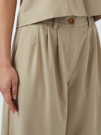 Lys beige bukser med folder, knaplukning og en glat tekstur. Stoffet ser let struktureret og skræddersyet ud.