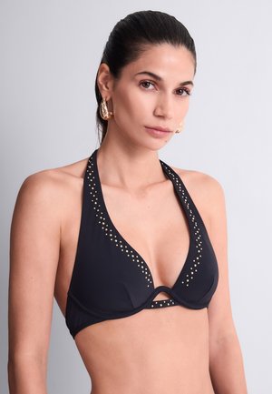 Femme aux cheveux foncés portant un haut de bikini halter noir orné de résille et des boucles d’oreilles anneaux dorés, posant devant un fond gris clair uni.