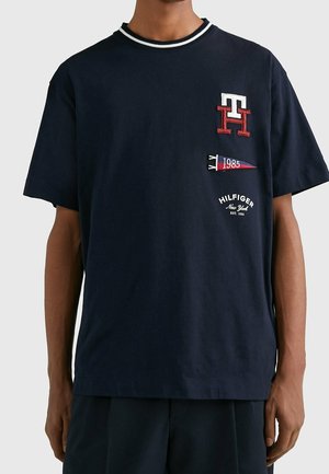 Marineblå T-shirt med hvidt og rødt "TH"-logo, et vimpel fra 1985 og teksten "Hilfiger New York Est. 1985" på brystet.