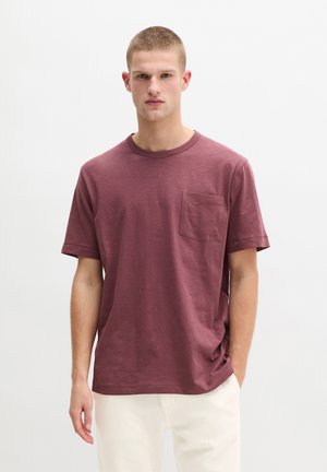 Junger Mann mit kurzen Haaren, der ein einfaches bordeauxrotes T-Shirt mit kurzen Ärmeln und Brusttasche sowie hellfarbene Hosen trägt, vor weißem Hintergrund steht.