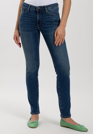 Slim-fit blue denim jeans with a slight fade, five-pocket design, and a button closure, paired with light green flat shoes.