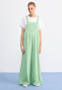 Groene katoenen overall met wijde pijpen, voorzien van verstelbare schouderbanden, een voorkeurszak en een gestructureerde stof. Gekozen boven een wit T-shirt.