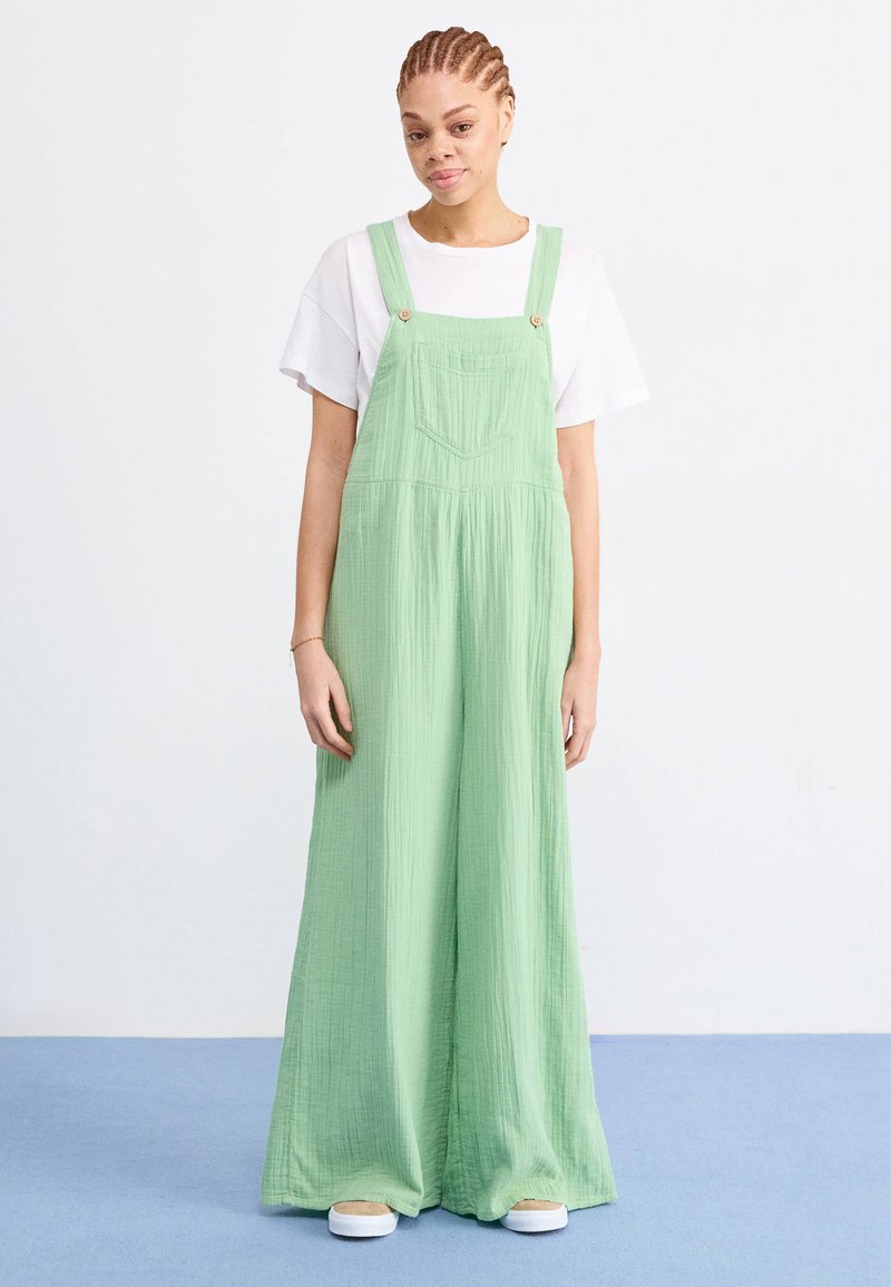 Groene katoenen overall met wijde pijpen, voorzien van verstelbare schouderbanden, een voorkeurszak en een gestructureerde stof. Gekozen boven een wit T-shirt.