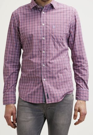 Mann trägt ein rot-blau kariertes langärmliges Hemd mit Knöpfen und graue Jeans, steht vor einem einfarbigen Hintergrund.
