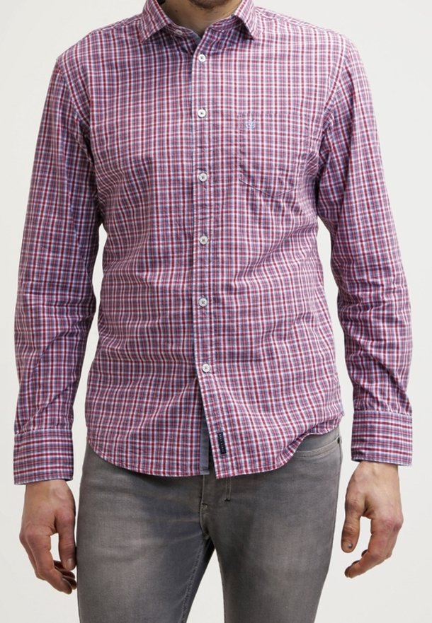 Homme portant une chemise à carreaux rouges et bleus à manches longues et un jean gris, debout devant un fond uni.