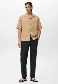 Una camicia beige a maniche corte con tasca, abbinata a pantaloni neri e sandali neri. La camicia presenta una vestibilità rilassata e un colletto a punta.
