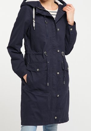 Femme portant un manteau à capuche bleu marine avec poches avant, fermetures à boutons et à fermeture éclair, et cordons de serrage ajustables à la taille, par-dessus un haut rayé.
