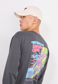 Young man in a beige cap and dark long-sleeve shirt with colorful climbing graphic and "The North Face" text on the back faces left.