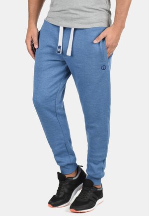 Pantalones joggers de algodón azul con cordones blancos, bolsillos laterales y detalle de logo. Combinados con zapatillas deportivas negras con un acento naranja.