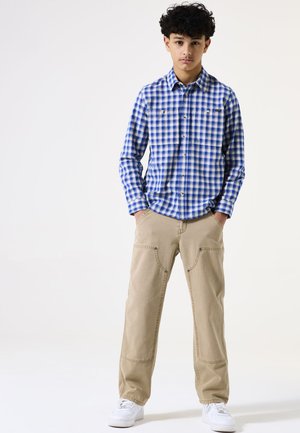 Camisa de manga larga a cuadros en azul y blanco, combinada con pantalones cargo beige y zapatillas blancas. Presenta dos bolsillos en el pecho y un corte relajado.