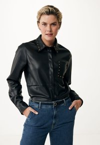 Camicia in ecopelle nera con borchie dorate sul colletto e sulla tasca, caratterizzata da una chiusura con bottoni e vestibilità slim. Abbinata a jeans in denim blu.