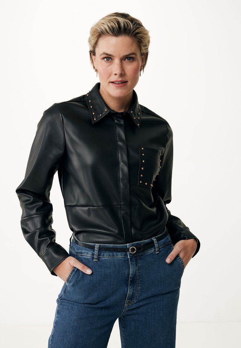 Camicia in ecopelle nera con borchie dorate sul colletto e sulla tasca, caratterizzata da una chiusura con bottoni e vestibilità slim. Abbinata a jeans in denim blu.