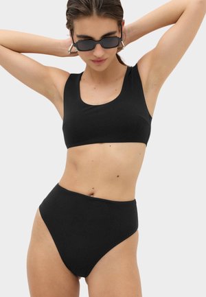 Kobieta w czarnym bikini z wysokim stanem, topem o kwadratowym dekolcie i prostokątnymi okularami przeciwsłonecznymi, pozująca z rękami za głową.
