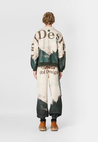 Sudadera de gran tamaño en color crema con estampado en verde oscuro y de montañas, combinada con pantalones sudadera a juego que presentan un logotipo y un patrón de paisaje, usados con botas color tostado.